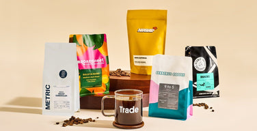 A glass coffee mug printed with the Trade logo, surrounded by bags from Metric, Broadsheet, Airship, Cerberus, and Atomic coffee roasters