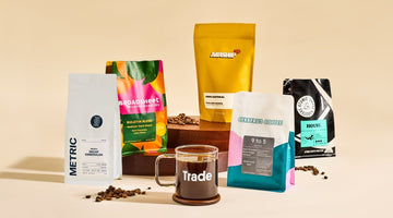 A glass coffee mug printed with the Trade logo, surrounded by bags from Metric, Broadsheet, Airship, Cerberus, and Atomic coffee roasters