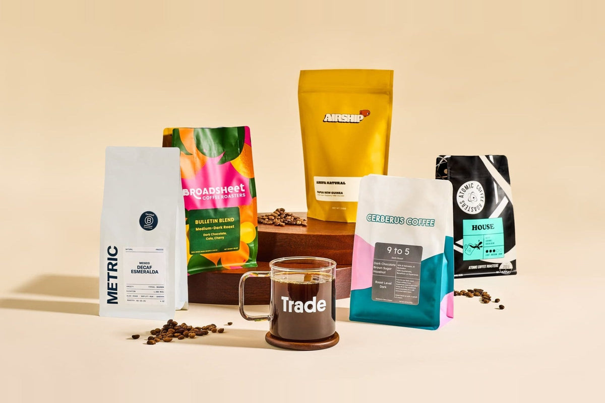 A glass coffee mug printed with the Trade logo, surrounded by bags from Metric, Broadsheet, Airship, Cerberus, and Atomic coffee roasters