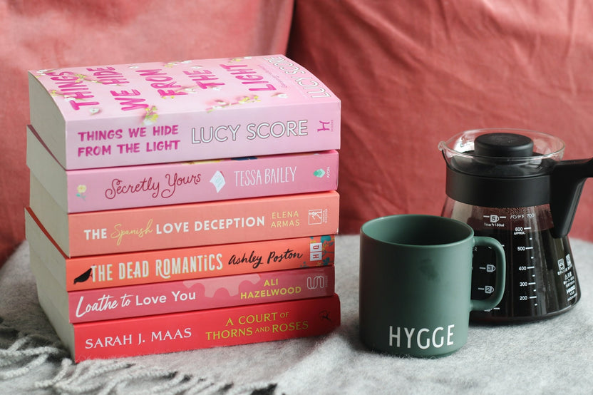 A cozy stack of pastel romance novels beside a coffee mug and French press, set against soft pink fabric for the perfect reading moment.