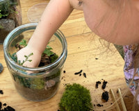 A picture of a young girl placing a plant inside and almost completed terrarium. There is another completed terrarium in the background.