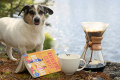 An orange romance book with coffe mug and a french press and a dog next to each other. In the backgound there is a lake.