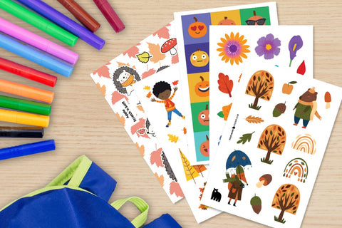 A selection of autumn themed stickers (pumpkins, flowers, trees) pictured on a desktop along with markers and a backpack.