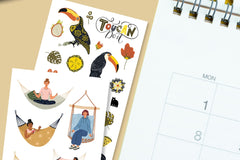 A spiral bound monthly calendar pictured alongside two sticker sheets.  One with women relaxing in hammocks and another with toucans.