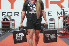 This image shows a very fit woman carrying two Gainz Boxes across a gym on weighted bars as part of her workout for the day.