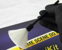 A blue box containing a forensikit is displayed in the back while a white fingerprint brush is held by a gloved hand in the foreground