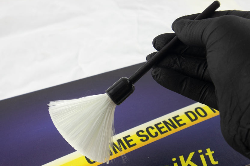A blue box containing a forensikit is displayed in the back while a white fingerprint brush is held by a gloved hand in the foreground