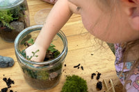 A close up view of a young girl placing a plant in a terrarium. There is moss dirt and rocks on the table she is using.