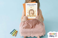 Girl age 2-5 in a light pink dress holding up her monthly Hooray Mail card with colorful crayons beside her.
