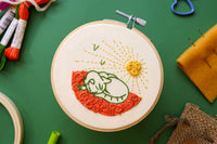 Embroidery project featuring a simple, green cat sleeping in a field of flowers and underneath birds and a bright sun.