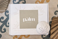 The Palm People box is sitting on a beach towel with a sand dollar and dried palm leaves. The box is decorated with sunshine prints.