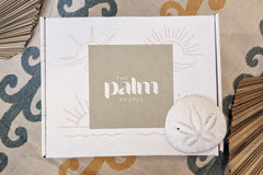 The Palm People box is sitting on a beach towel with a sand dollar and dried palm leaves. The box is decorated with sunshine prints.