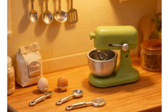 This is a photograph of a tiny miniature kitchen set.  There are eggs and flour and a green stand mixer. 