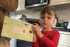 A boy in the kitchen holds up the green envelope for the letter he is mailing back to Zip, decorated with heart and dinosaur stickers.