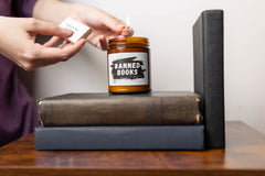 A candle named Banned Books sits atop a stack of books. It is being lit by a person holding a lit match and a box of matches.