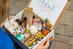 An over the shoulder view of an unboxing. This box showcases colored pencils, a game, facial cream and soap.