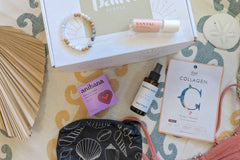 These Palm People box products are displayed on a beach towel: shower steamer, bracelet, small bag, tanner, perfume, and face mask.