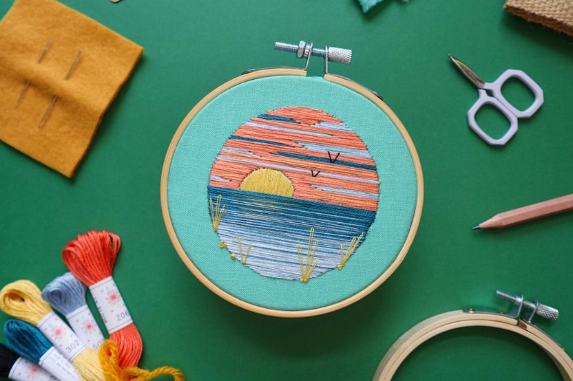 Embroidery project featuring a colorful sunrise or sunset and surrounded by various embroidery kit supplies.