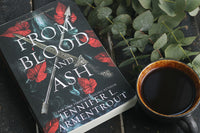 High quality close up photo of a book called From Blood and Ash that is a romance book with a coffe mug and leaf next to is.