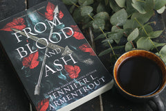 High quality close up photo of a book called From Blood and Ash that is a romance book with a coffe mug and leaf next to is.