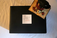 This picture shows the black box with the logo and a raven, rose, and skull statue on a light and shaded background