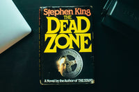 Close up image of a simple one book called the dead zone from an author named Stephen King with a macbook next to it.