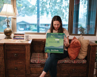 A woman sitting at a book nook while looking through a Box of Books subscription box with a smile on her face. 