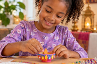 A child decorates a colorful Diwali diya with bright foam shapes and gems, illustrating the hands-on holiday craft 