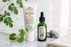 Queen City Skincare soothing facial cleansing foam and calming facial serum displayed on marble with green leaves, spa stones, and a folded 