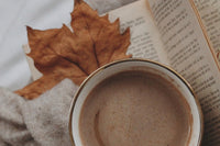 AN OPEN BOOK SITS UNDER A BROWNING LEAF AND A CUP OF CAPPUCINO. A SOFT SWEATER LIES ALONGSIDE TO COMEPLETE THE COZY IMAGE.