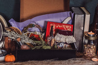 opened black box filled with a book, socks, an engraved cooking spoon, dried herbs and sitting next to candles and more dried herbs