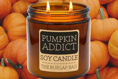 Pumpkin Addict 16oz double-wick soy candle by The Burlap Bag, spiced pumpkin scented amber jar against small orange pumpkins