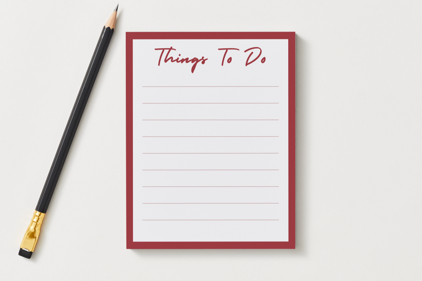Things To Do notepad with a deep red border and lined pages, shown next to a black pencil on a soft gray desk