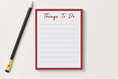 Things To Do notepad with a deep red border and lined pages, shown next to a black pencil on a soft gray desk