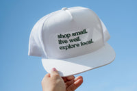 A hand holding a white cap with green text saying "shop small, live well, explore local" for the Explore Local Box monthly subscription
