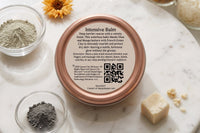 Top-down view of a Queen City Skincare Intensive Balm tin on a marble tray, surrounded by green clay, botanical ingredients, oils, waxes, an
