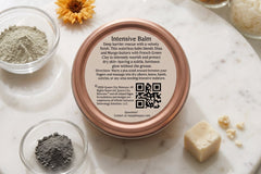 Top-down view of a Queen City Skincare Intensive Balm tin on a marble tray, surrounded by green clay, botanical ingredients, oils, waxes, an