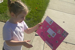 A young child on the sidewalk studies her pink Zip's Mailbox Club envelope, covered in stickers and real postage stamps, before opening it.