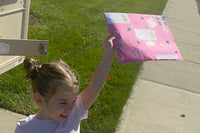 A young child at the family mailbox lifts her pink Zip's Mailbox Club envelope high in the air on mail day, grinning in the sunshine.
