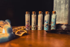 a photo of 5 jars of bath salts mixed with dried florals and herbs. set in candlelight and next to a feather and dried orange slices.