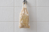 The Turmeric & Black Pepper bar inside the sisal soap saver bag, hanging from a hook on a wet white-tiled shower wall to drain