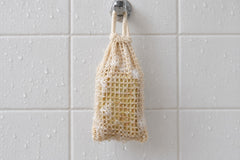 The Turmeric & Black Pepper bar inside the sisal soap saver bag, hanging from a hook on a wet white-tiled shower wall to drain