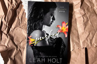 A slender woman holding a shaw against her body. The title Splashed appears in the middle. Flanked by two flowers. 