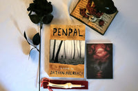This is a picture of the book titled Penpal with a scary moon picture, black rose, bone pen with red pen holder, and raven statue
