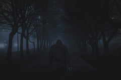 Dark and scary photo of a person walking down the road with many trees next to him and infront of him and cars in the corner.