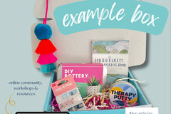 A picture of an example box featuring a bright coloured Pompom tassel, DIY pottery kit, therapy putty, a colouring book, page markers & info
