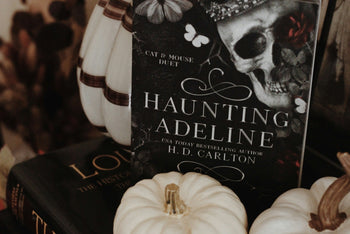 High quality image of horror books called haunting adeline from h d carlton stacked on each other with horror items around it.