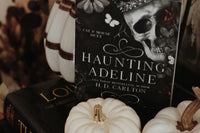 High quality image of horror books called haunting adeline from h d carlton stacked on each other with horror items around it.