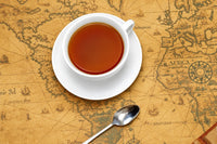 Image featuring a white tea cup with tea in it and saucer with a silver spoon on an old brown antique map. 
