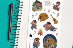 A notebook and pen pictured with a sticker sheet featuring a camping bear, singing fireside with his guitar and walking through the forest